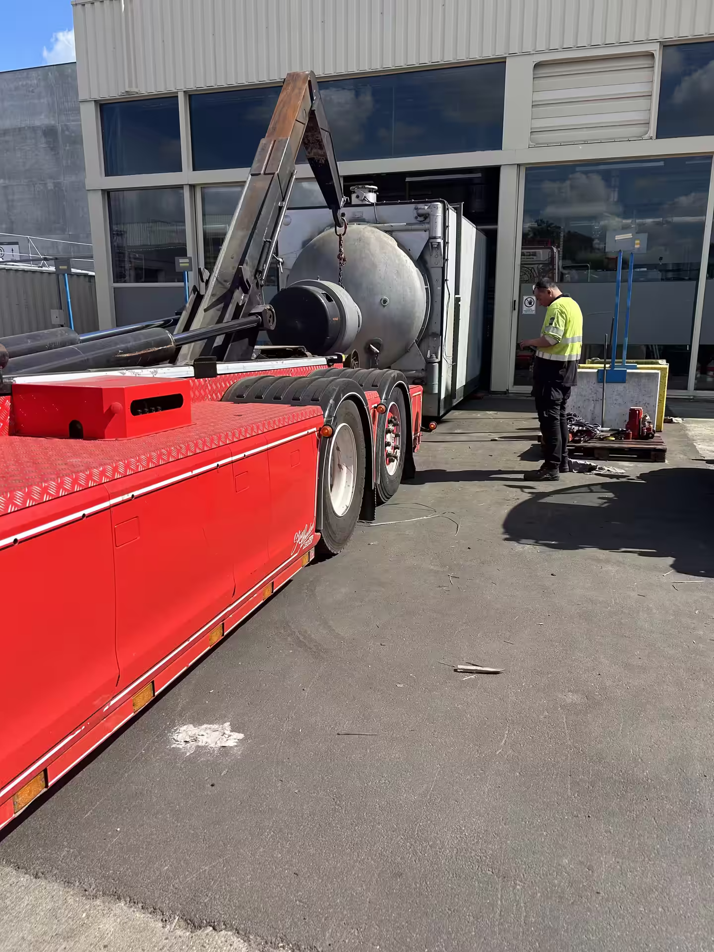 Grote metalen ketel wordt met de haakarm op een rode vrachtwagen geladen bij een bedrijfspand