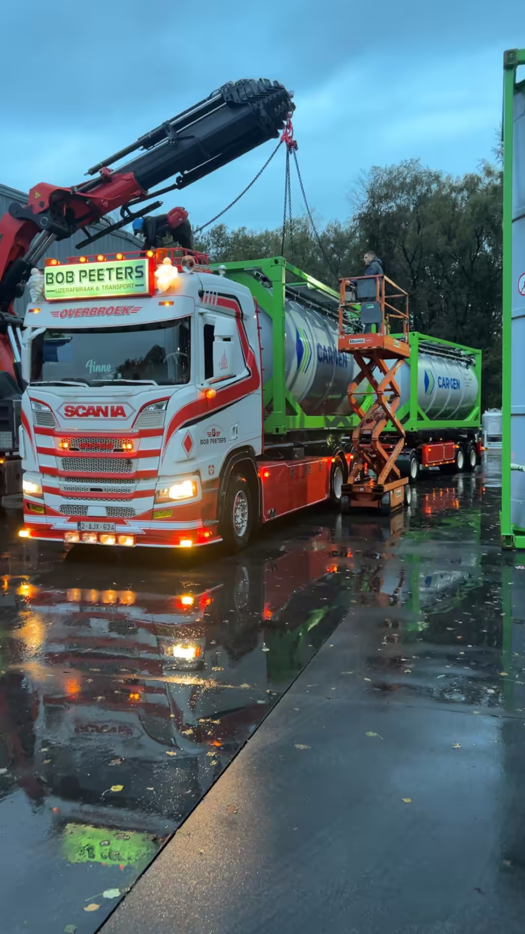 Scania-kraanwagen van Bob Peeters laadt tankcontainers in de schemering, met een medewerker op een schaarlift