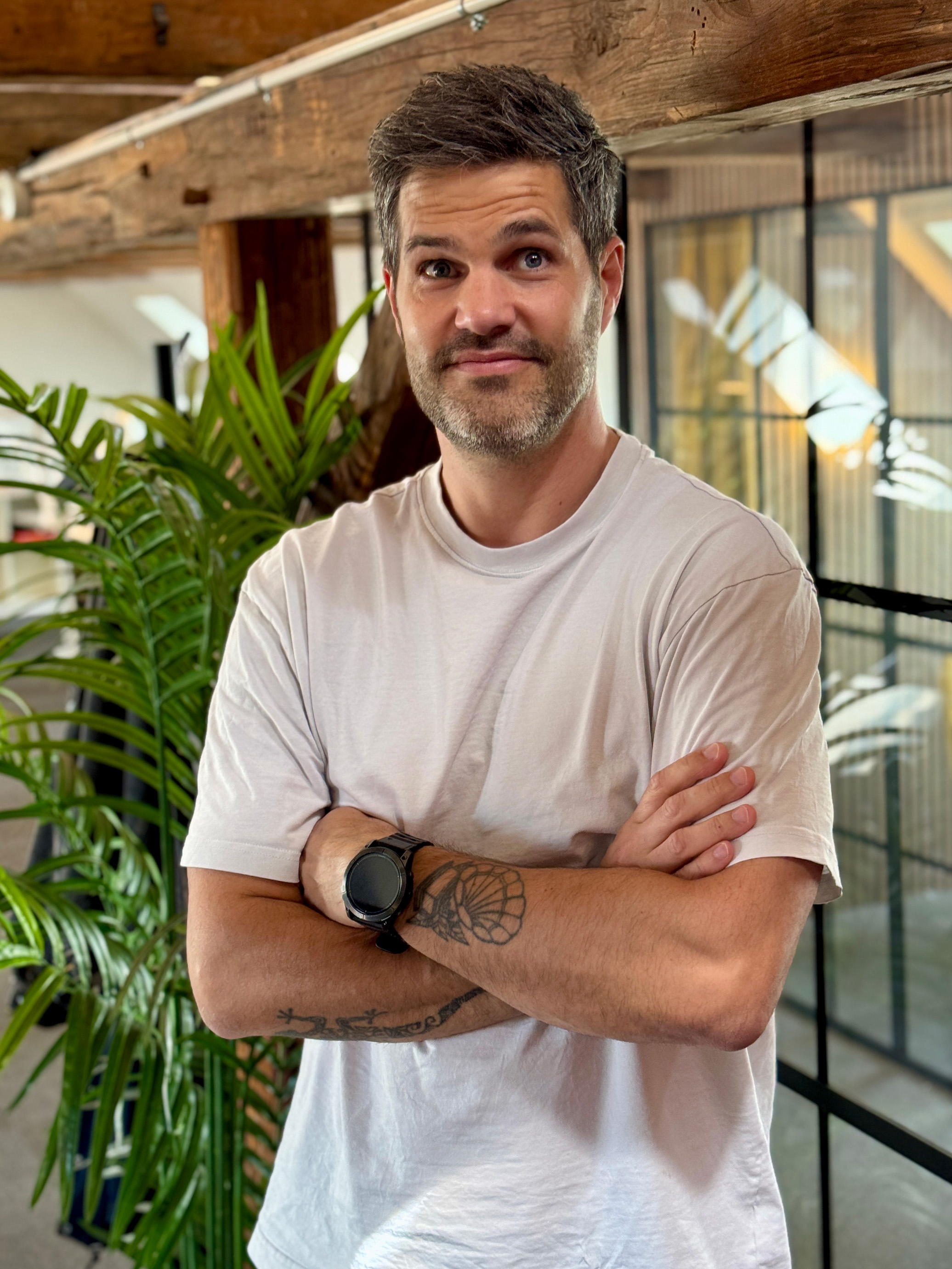 Mand med kort hår og skæg med armslagte tatoveringer og sort ur i lys T-shirt indendørs.