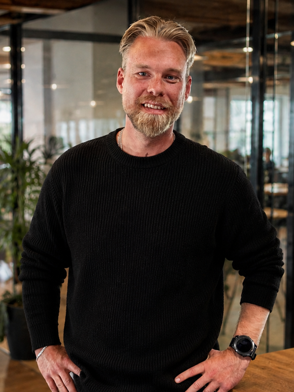 Smilende mand med lyst hår og skæg iført sort sweater i et moderne kontormiljø.