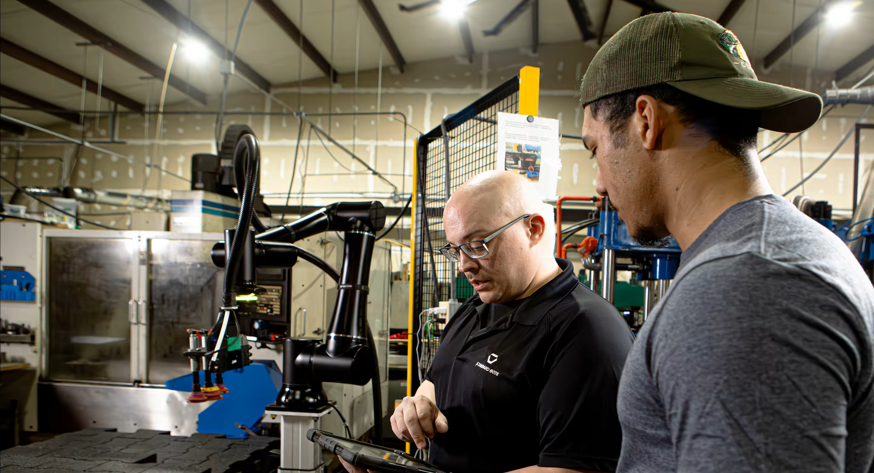 Two operators reviewing a tablet beside a Standard Bots robot arm tending a CNC machine