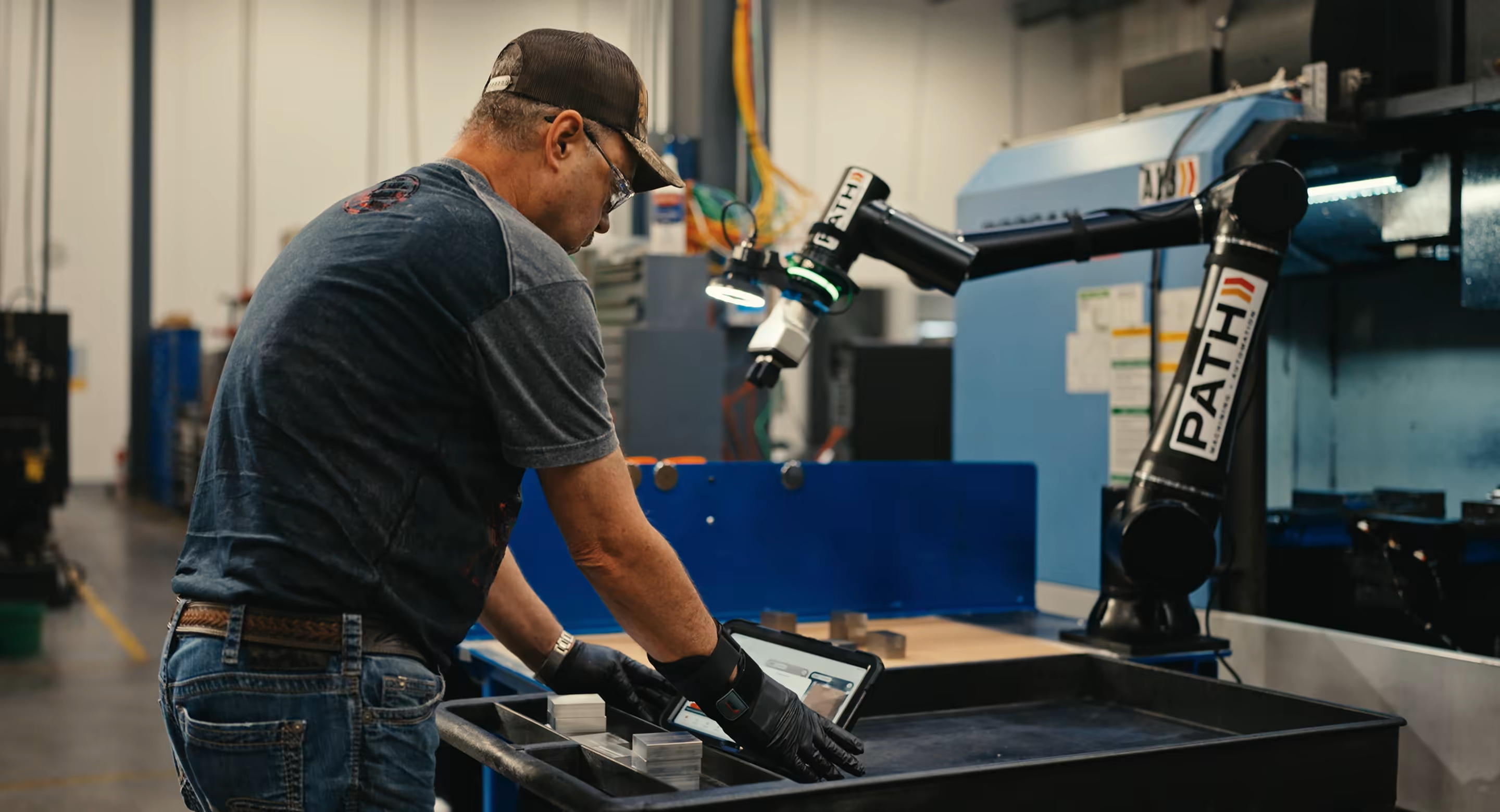 An operator loading parts at a workstation while a Standard Bots robot arm tends a drill press