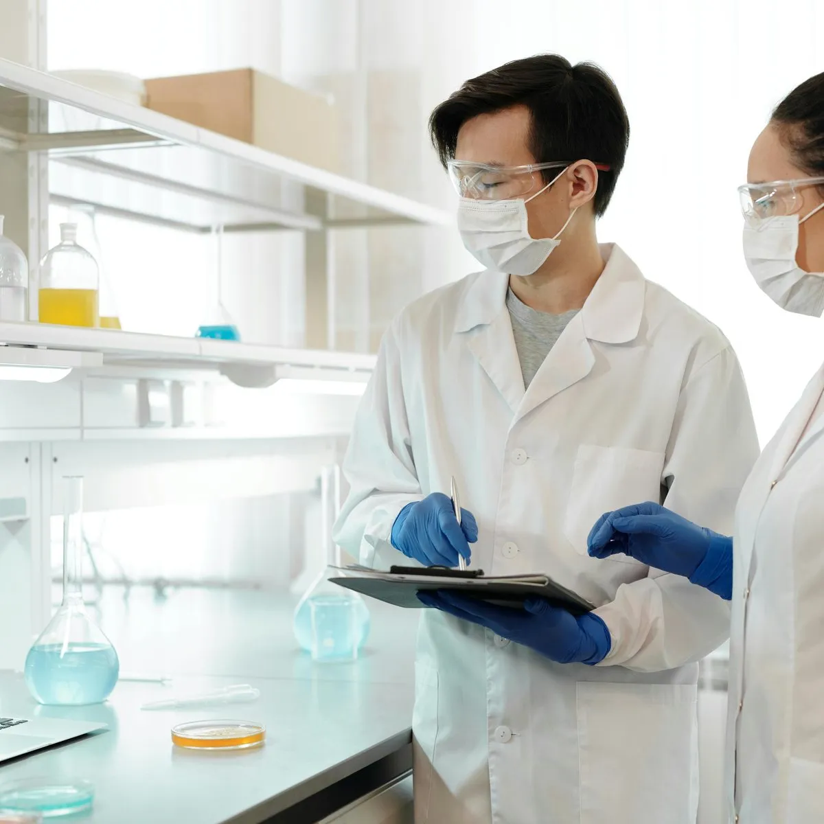 Two lab technicians in safety glasses reviewing test records on a clipboard