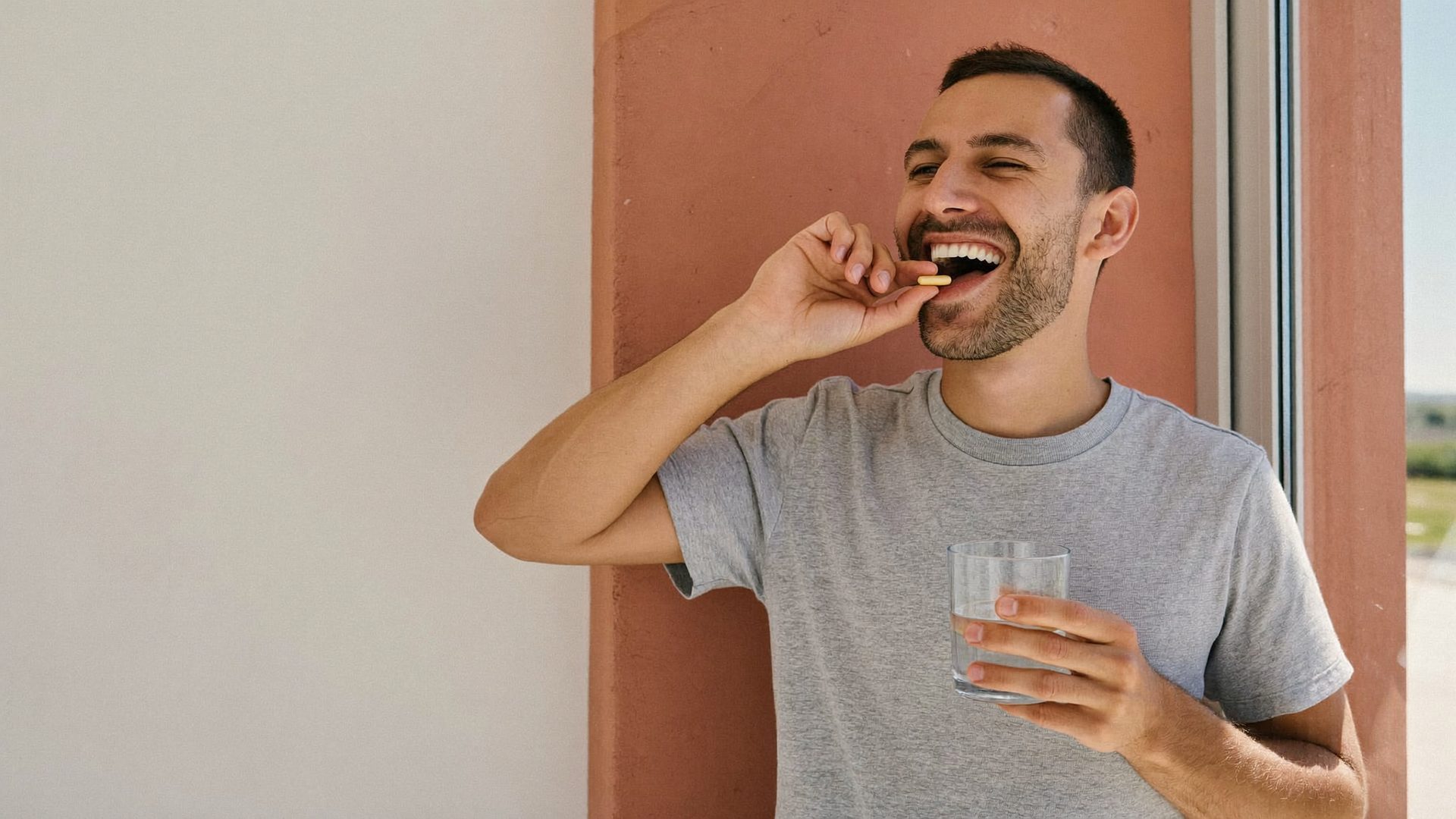 Man taking a capsule with a glass of water by a sunlit window