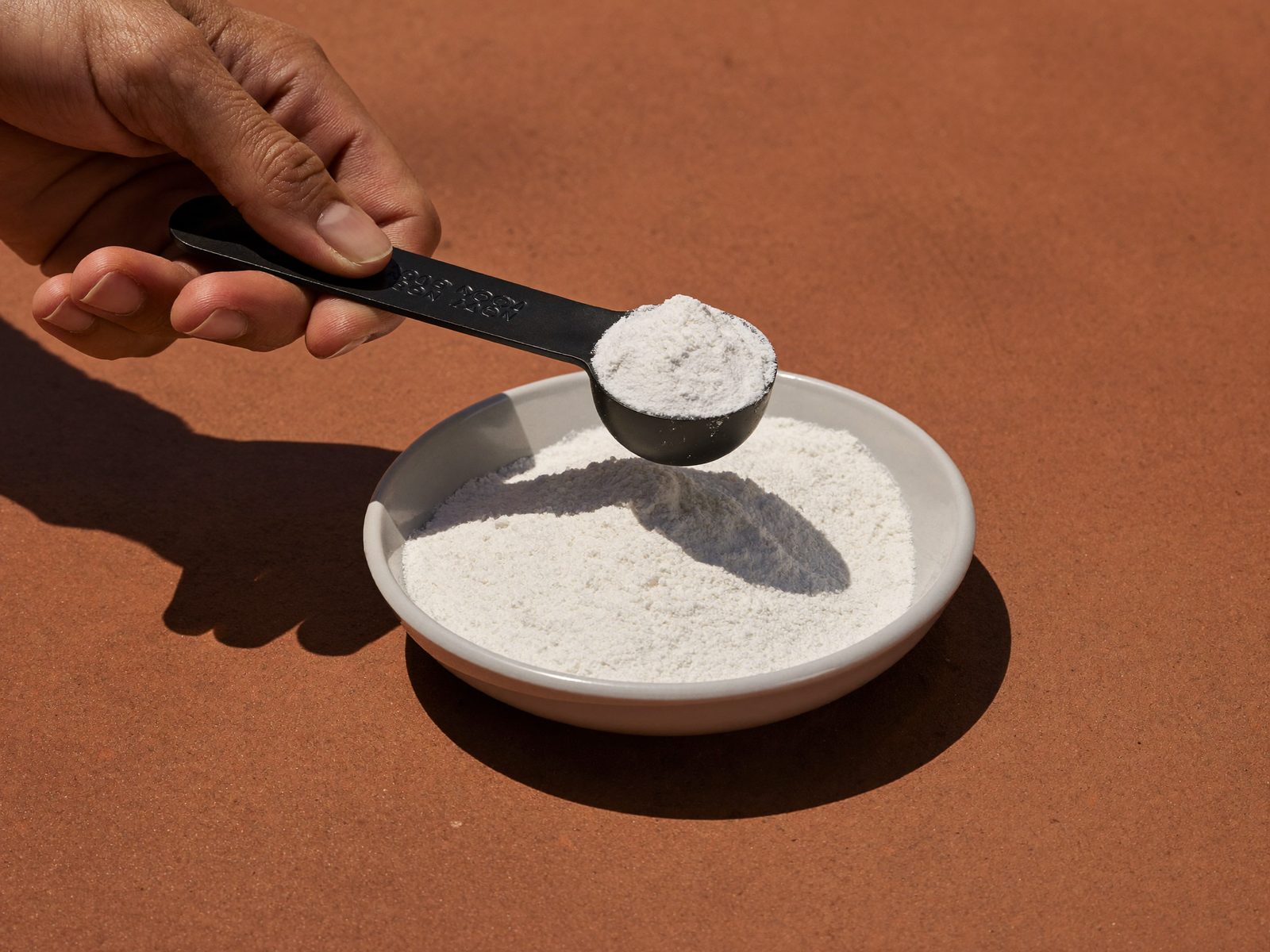 Hand lifting a scoop of white supplement powder from a dish