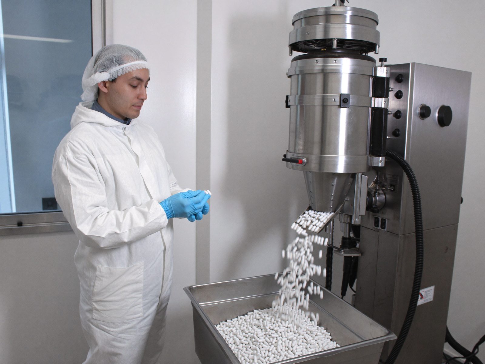 Technician in a cleanroom gown checking white capsules discharging from a capsule filling machine