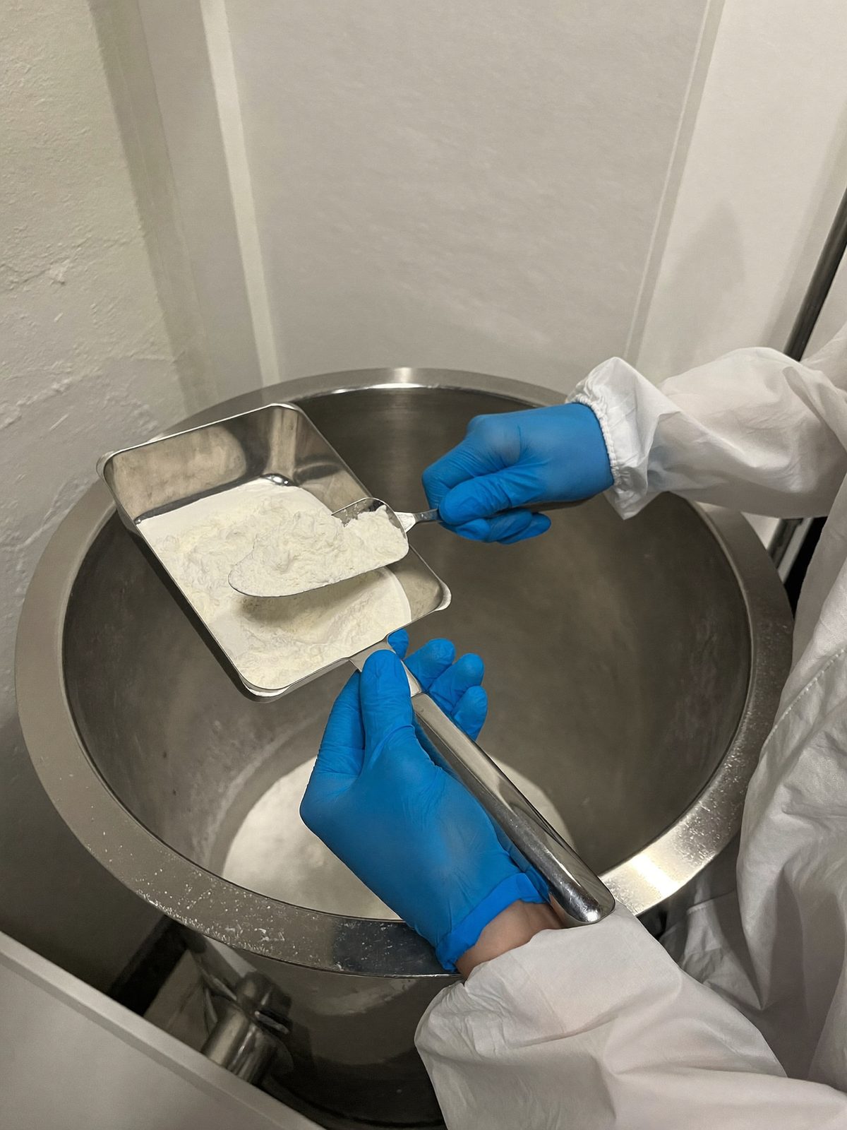 Gloved hands taking a scoop sample of white powder from a stainless drum