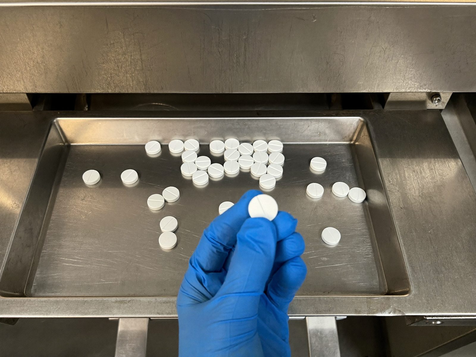 Gloved hand checking a round white tablet at the discharge tray of the tablet press