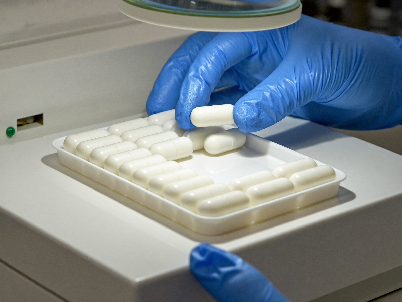 Technician checking a tray of white capsules under a magnifier lamp