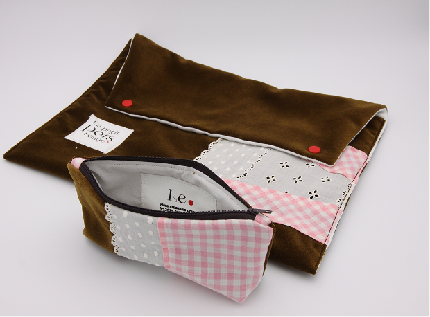 A brown fabric pouch with red snap buttons and a patchwork design featuring pink gingham, gray with white polka dots, and white fabric with small black floral embroidery, accompanied by a matching smaller zippered pouch with lace trim and a visible label inside.