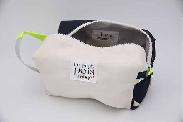 A small rectangular canvas pouch with a white body, navy sides, and a white zipper partially open showing a tag inside; the front has a sewn label reading 'Le petit pois rouge' and the pouch has a side loop with neon yellow and gray fabric.