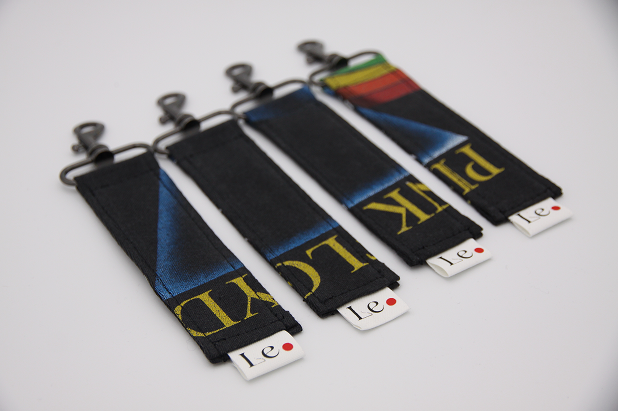 Four fabric keychains with black backgrounds and yellow text, each attached to a metal clip; the first two display partial letters 'CLKD' and 'LOCK', the third and fourth have a partial rainbow stripe design, laid out side by side on a white surface.