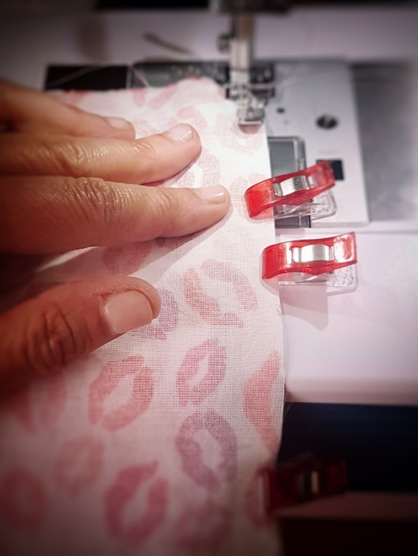 Close-up of a hand guiding pink fabric with lip print pattern through a sewing machine, secured with red clips.