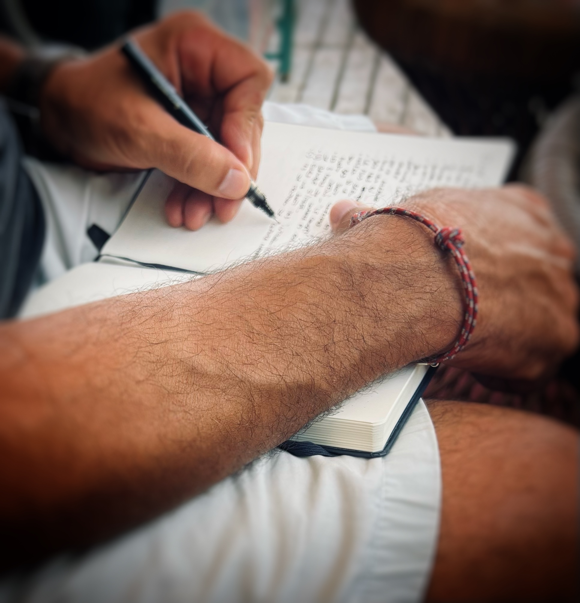 Close-up of a person writing in a notebook with a pen, showing the forearm with a red and gray woven bracelet.