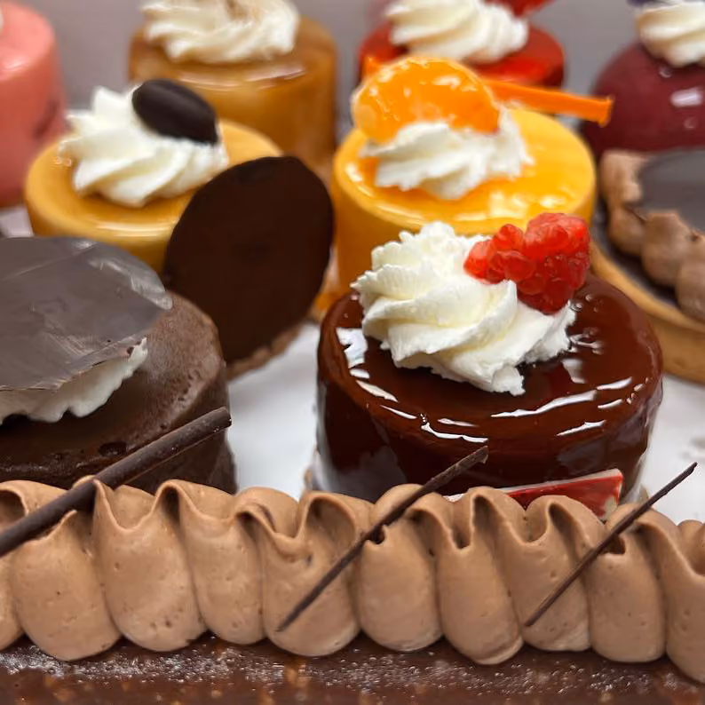 Assorted small pastries topped with whipped cream, fruit, and chocolate decorations on display.