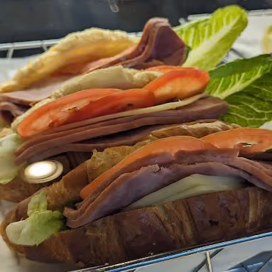 Two croissant sandwiches with slices of ham, tomato, cheese, and lettuce on a metal rack.