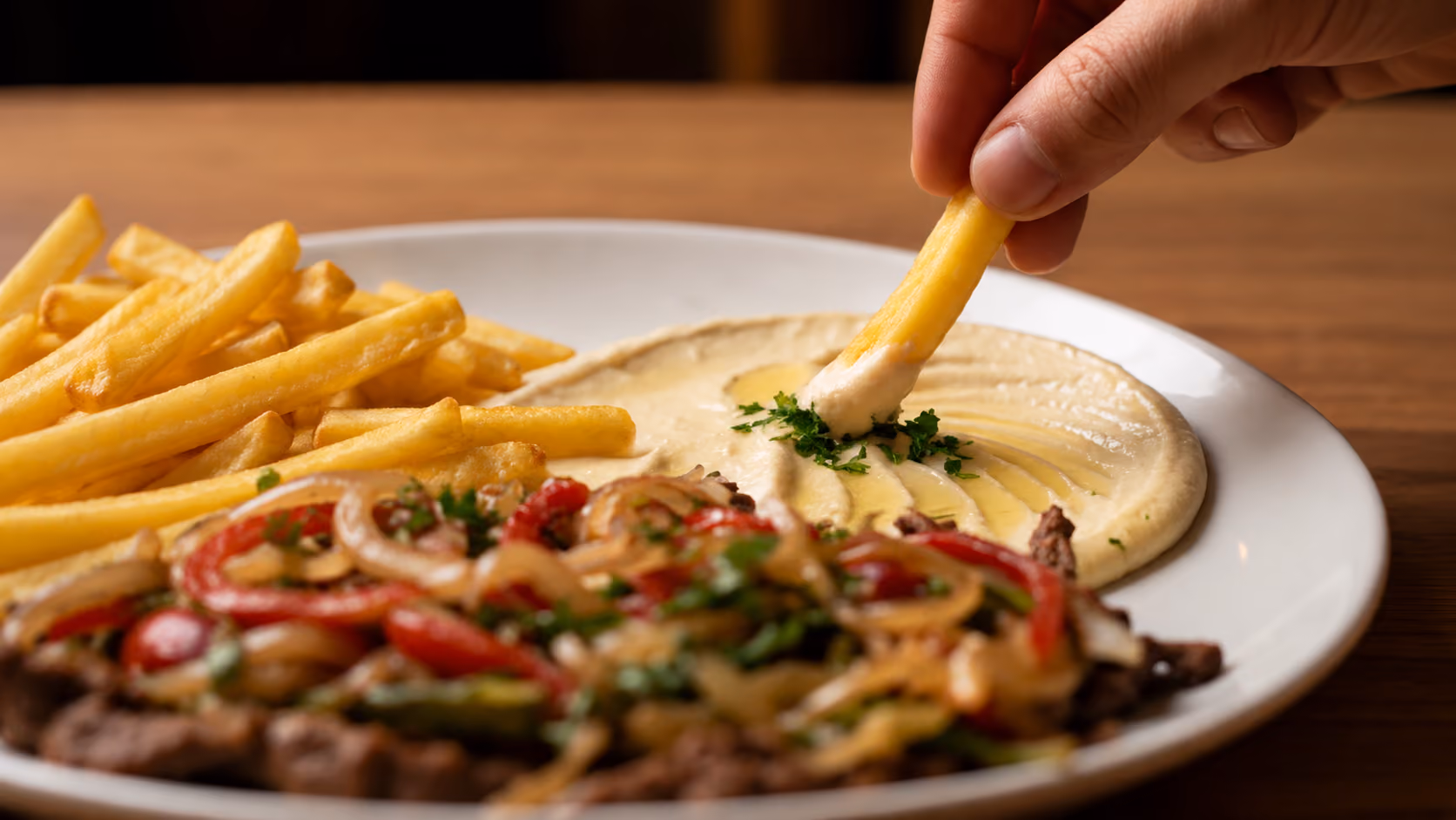 Hand dipping a French fry into hummus on a plate with fries and grilled vegetables.