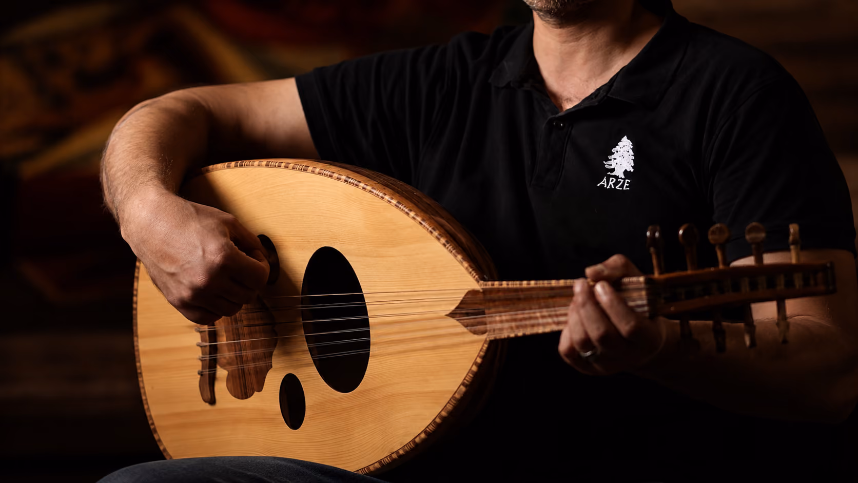 Person playing a wooden oud string instrument, wearing a black shirt with a white tree logo.