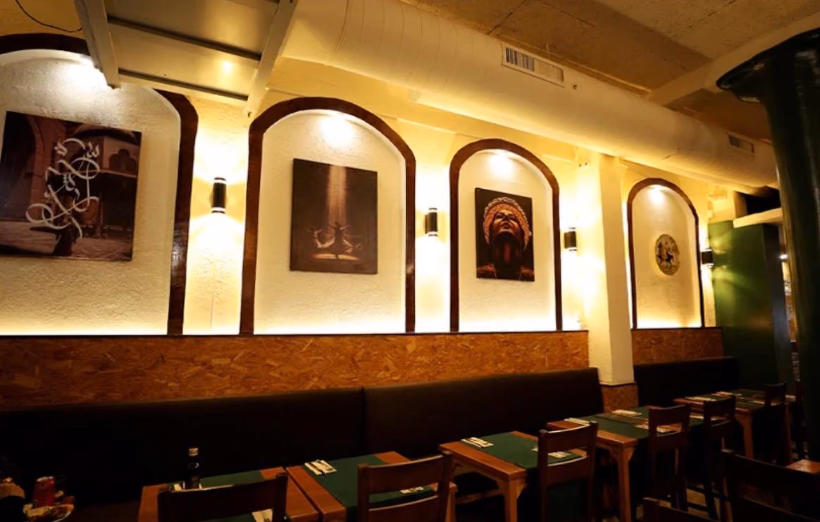 Cozy restaurant interior with dark wooden tables, chairs, cushioned bench, and illuminated wall art.