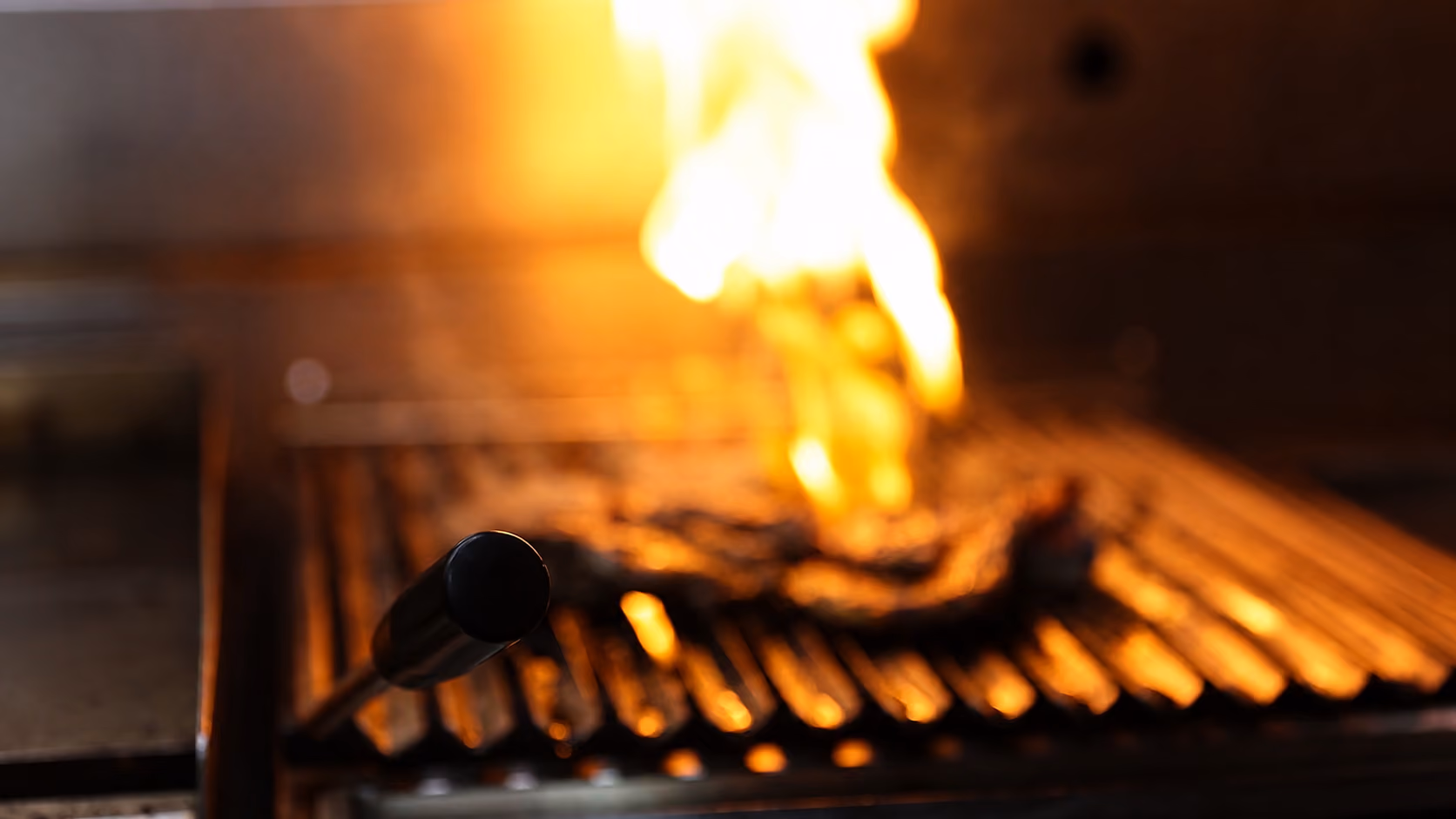 Close-up of a grill handle with flames and glowing grill grates in the background.