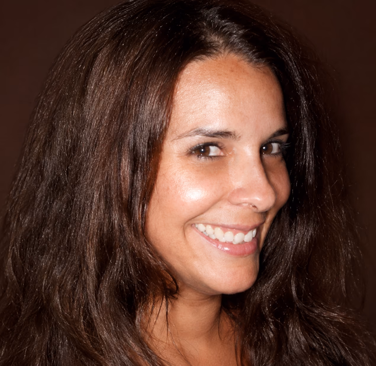 Smiling woman with long brown hair and brown eyes looking to the side against a dark background.