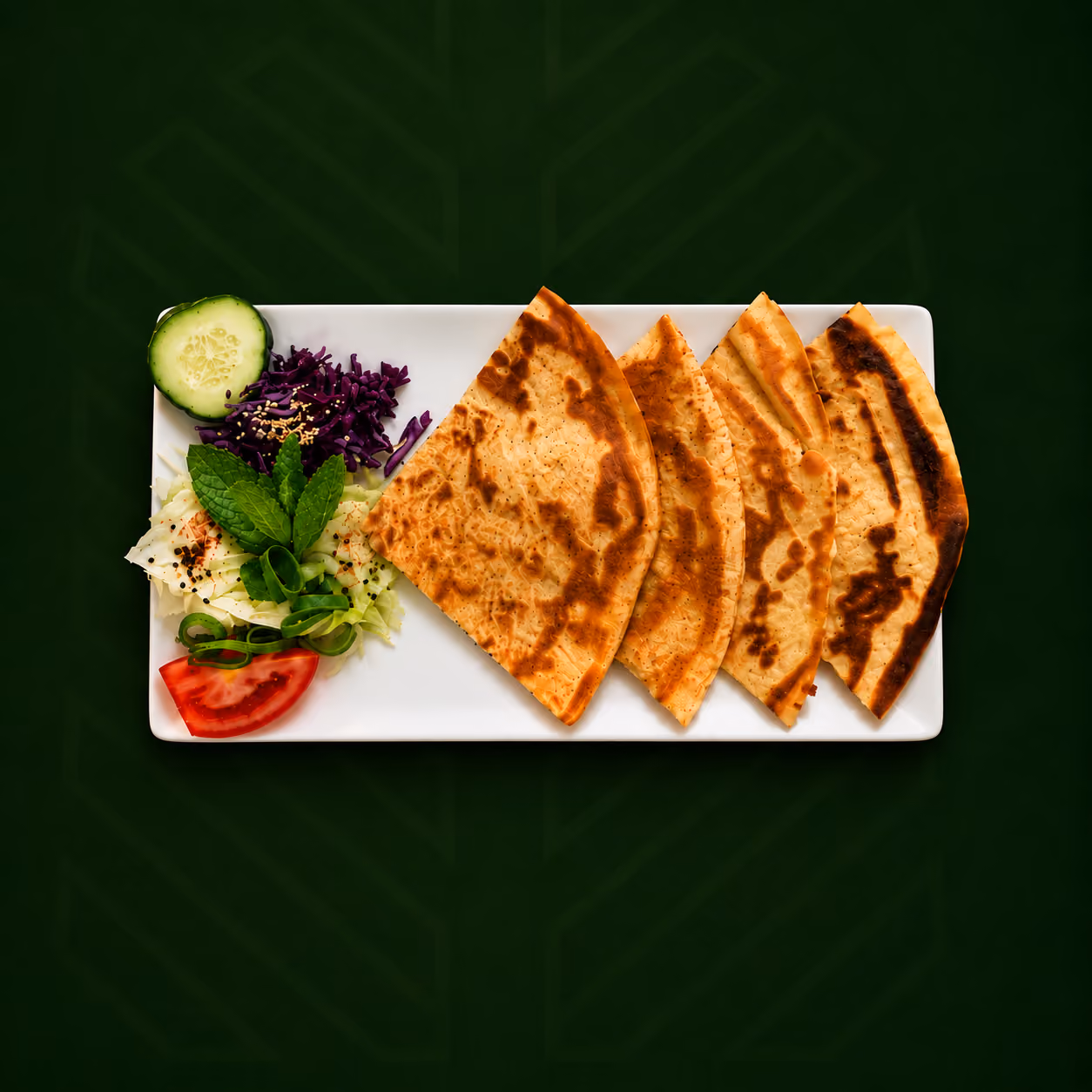 Rectangular white plate with four folded pieces of grilled flatbread arranged in a row, accompanied by a garnish of sliced cucumber, shredded purple cabbage, sliced tomato, shredded lettuce with black sesame seeds, chopped green onions, and fresh mint leaves on a dark green background.