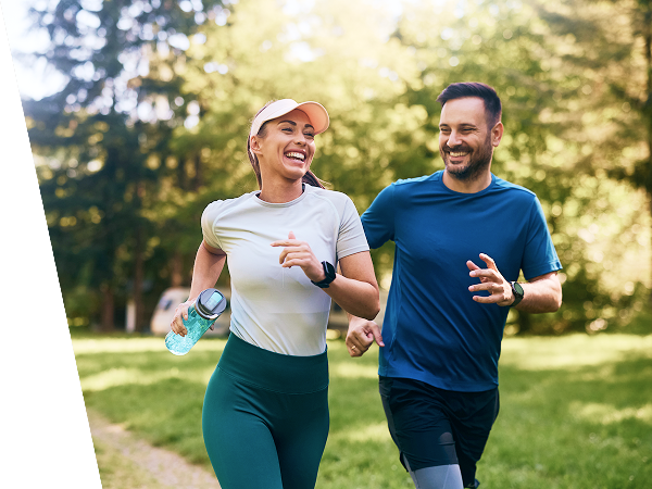 Smiling man and woman jogging together in a sunny park with green trees and grass.