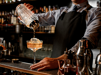 Bartender in apron pouring a cocktail from a shaker into a coupe glass at a bar.
