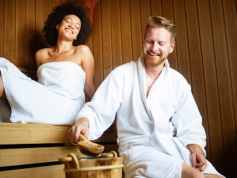 Smiling man and woman relaxing in a wooden sauna, wearing white towels and robes.