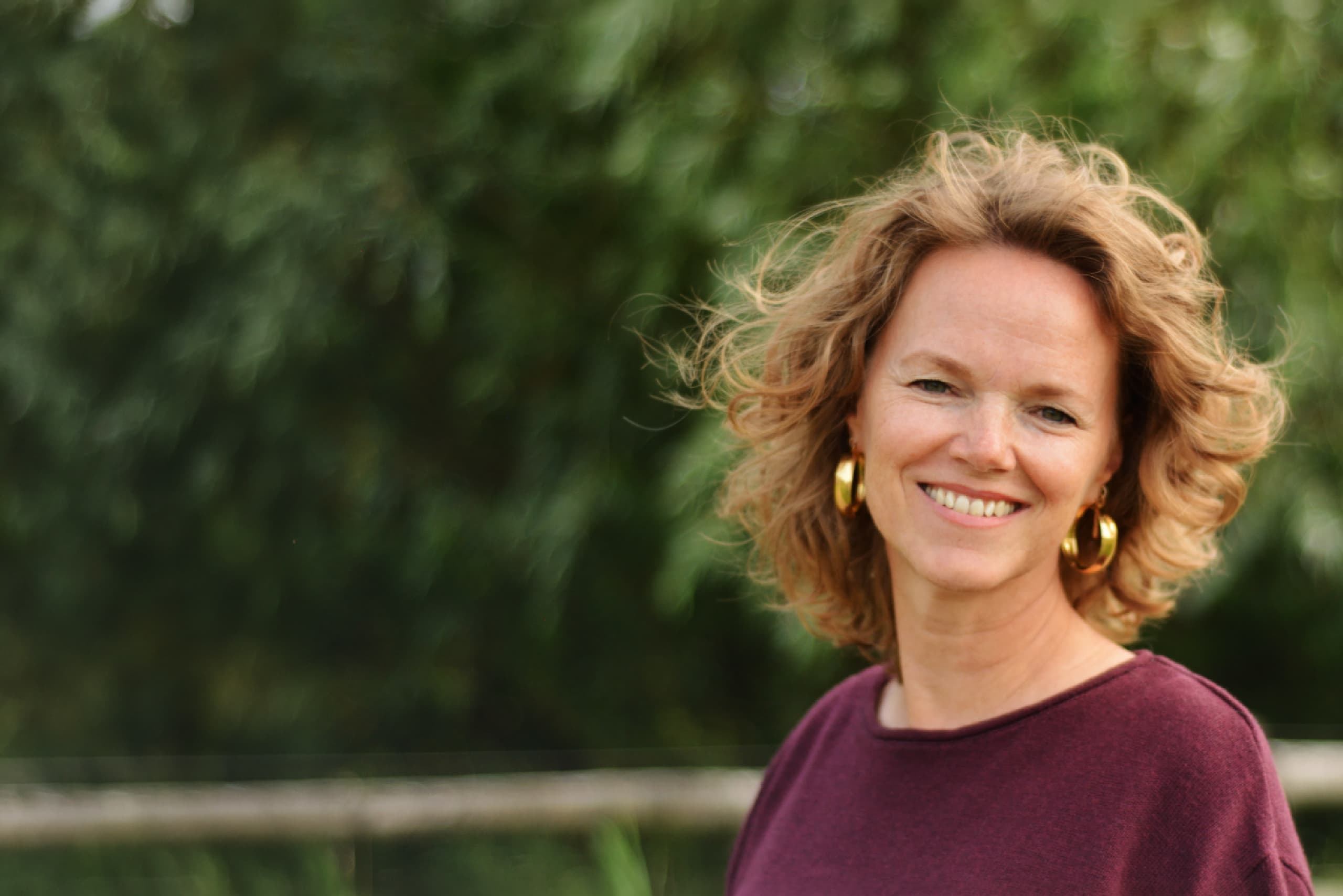 Vrouw met golvend lichtbruin haar, gouden oorbellen en een bordeauxrode trui lacht naar de camera met een wazige groene achtergrond.