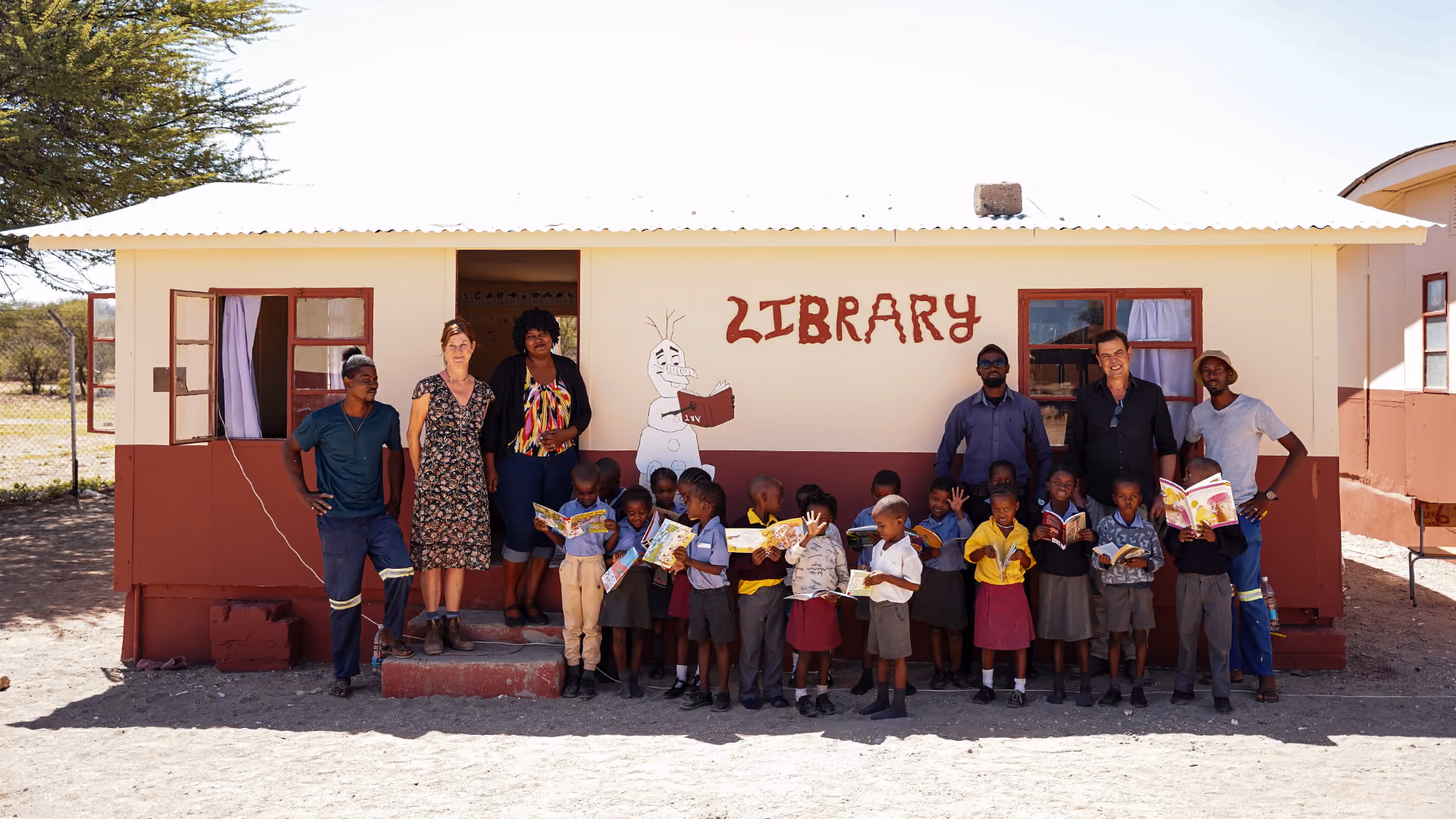 Eine Gruppe von Erwachsenen und Schulkindern posiert stolz mit Büchern vor einem kleinen, hellgelben Bibliotheksgebäude mit der Aufschrift „LIBRARY“.