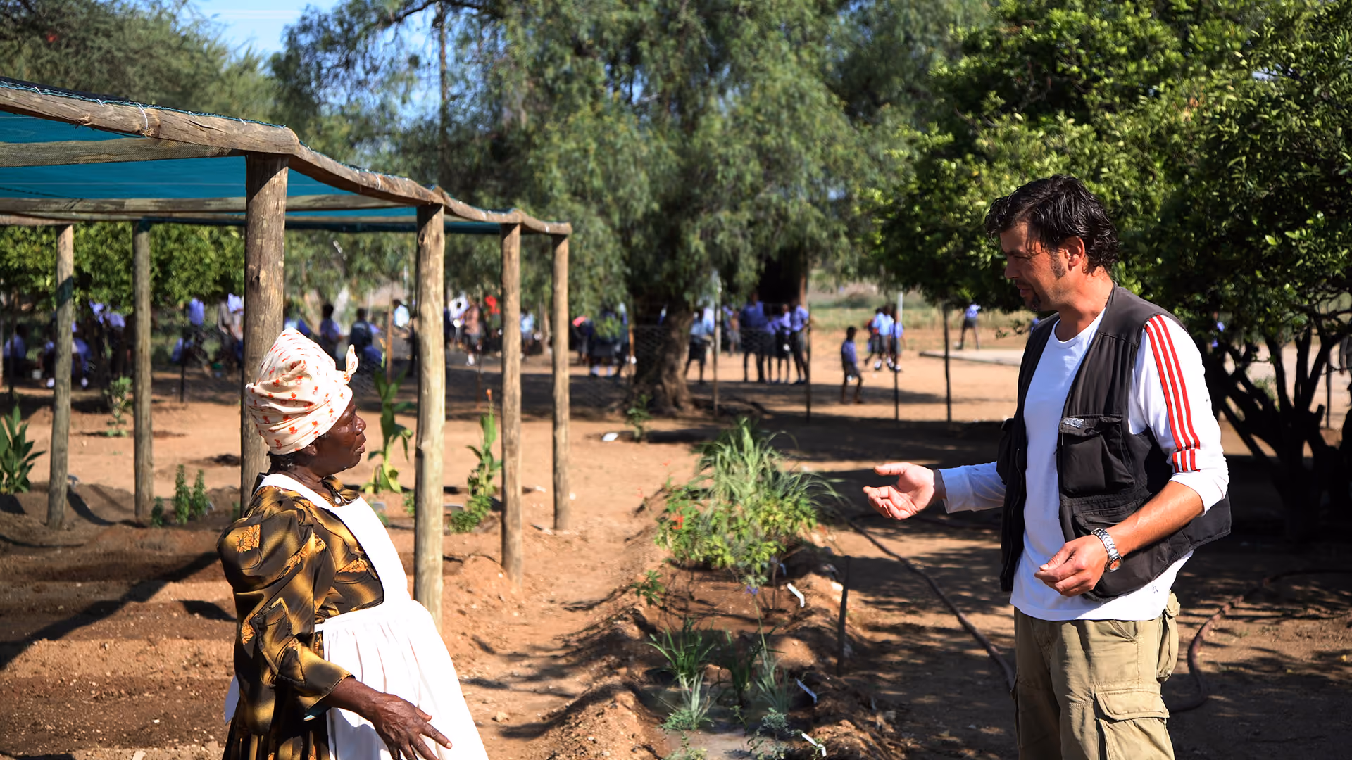 Eine Frau in traditioneller Kleidung unterhält sich in einem Garten mit einem Mann, während im Hintergrund Schulkinder zu sehen sind.