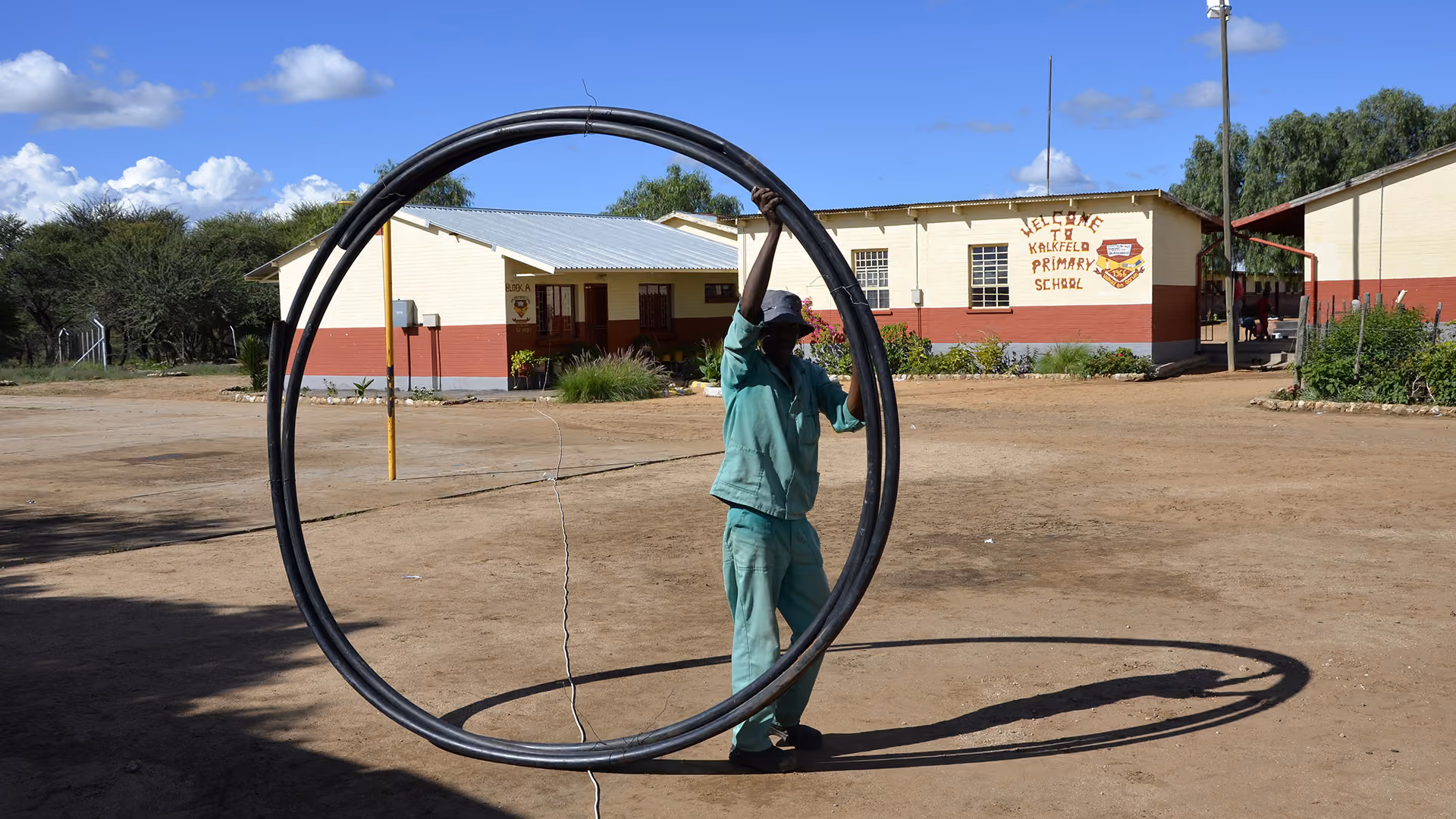 Ein Arbeiter in grüner Kleidung balanciert einen riesigen Ring aus schwarzen Wasserrohren auf dem sandigen Hof vor der Kalkfeld Primary School.
