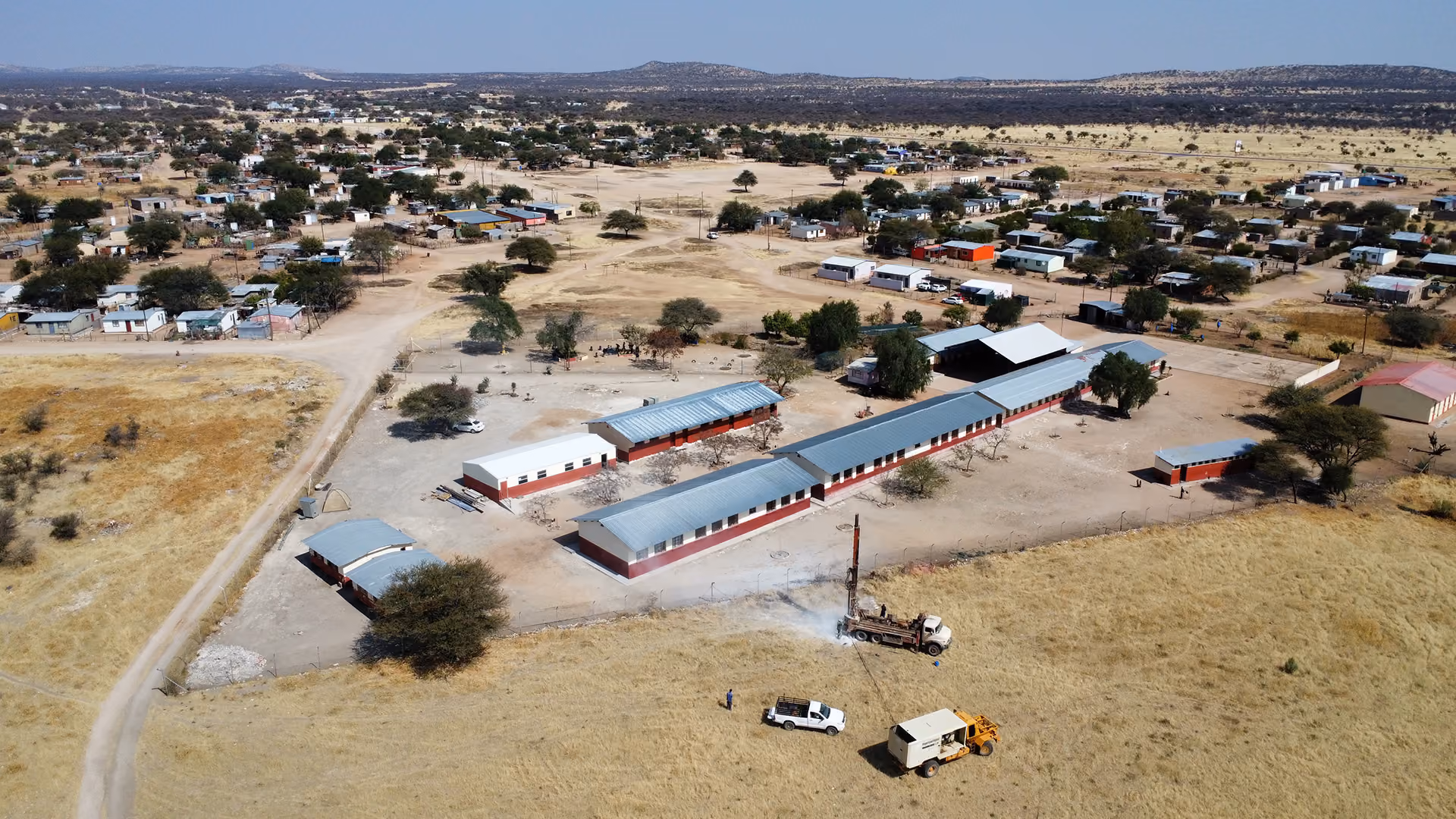 Eine Luftaufnahme zeigt einen Schulkomplex aus mehreren einstöckigen Gebäuden in einer weiten, trockenen Landschaft. Ein Brunnenbohrgerät ist auf einem nahegelegenen Feld im Einsatz, umgeben von zwei Pick-ups und einem gelben Lkw. Im Hintergrund ist eine verstreute ländliche Siedlung zu sehen.