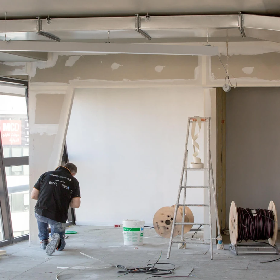 Worker preparing a wall in an office under renovation with ladder and cable reels