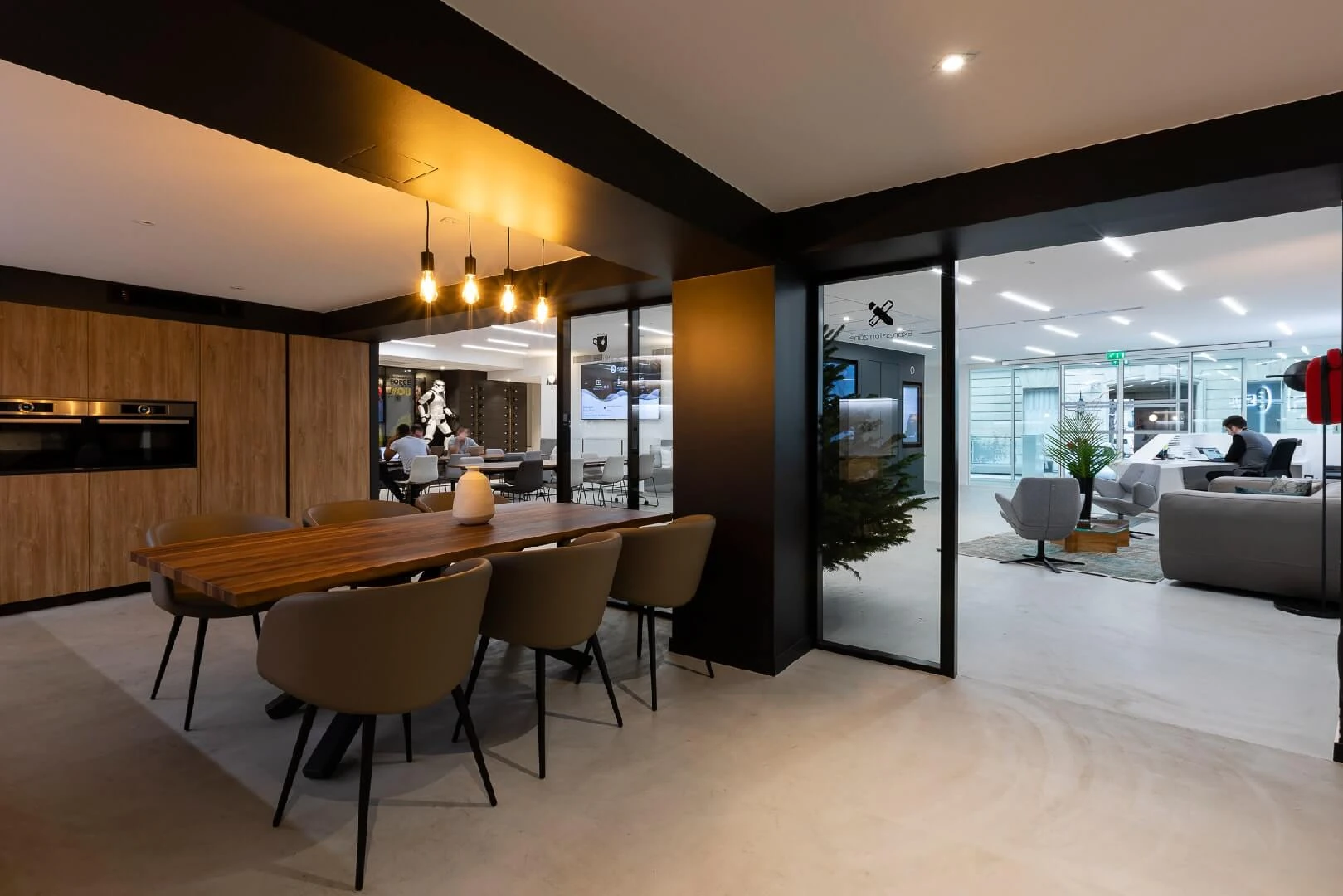 Office kitchen with dark wood cabinetry and a long dining table next to a glass-walled lounge