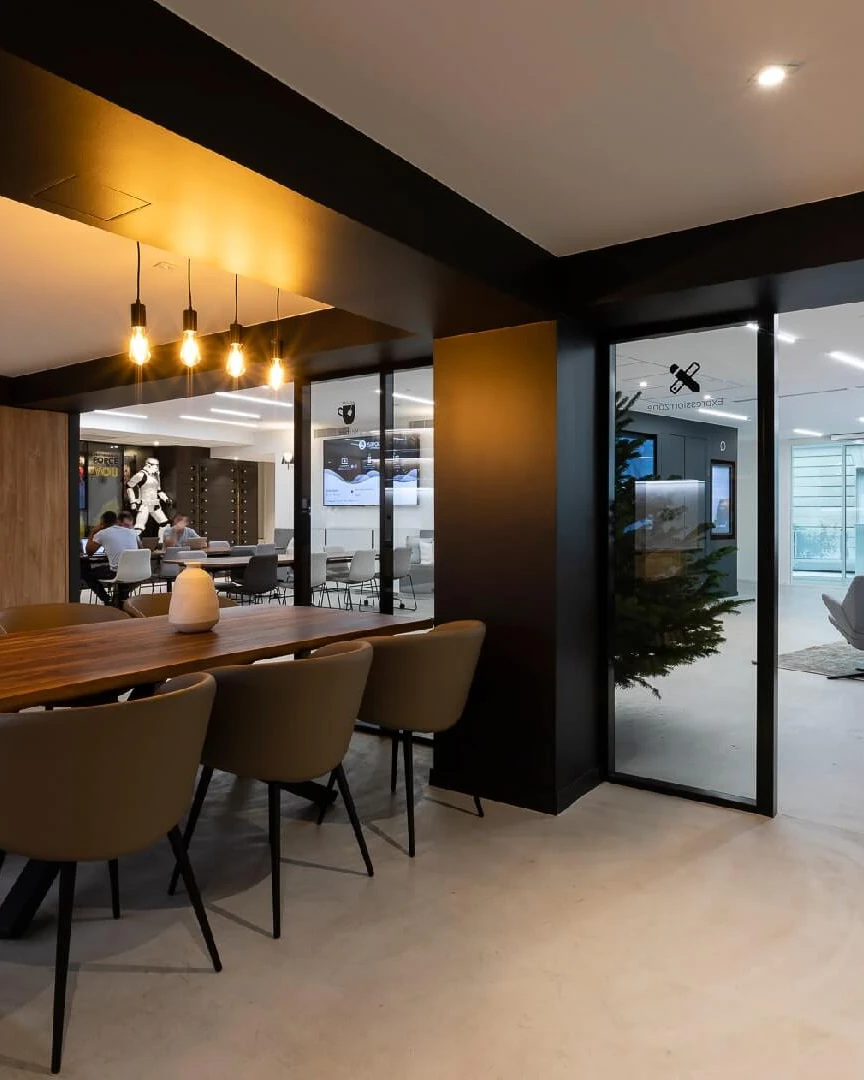 Office kitchen with dark wood cabinetry and a long dining table next to a glass-walled lounge