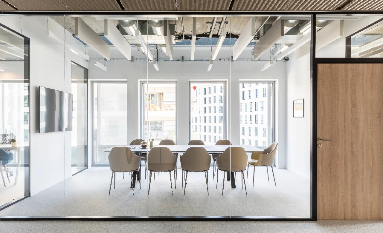 Glass-walled meeting room with a long table and beige chairs under an exposed ceiling with wooden slats