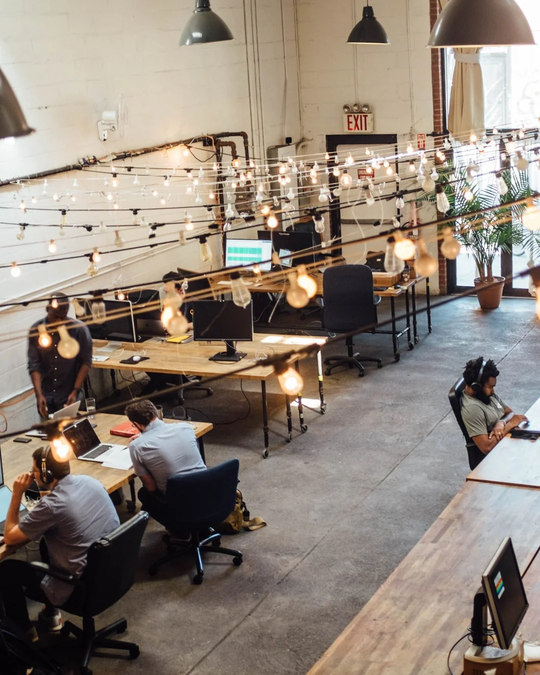 Open-plan office with long wooden desks, string lights and people working at computers