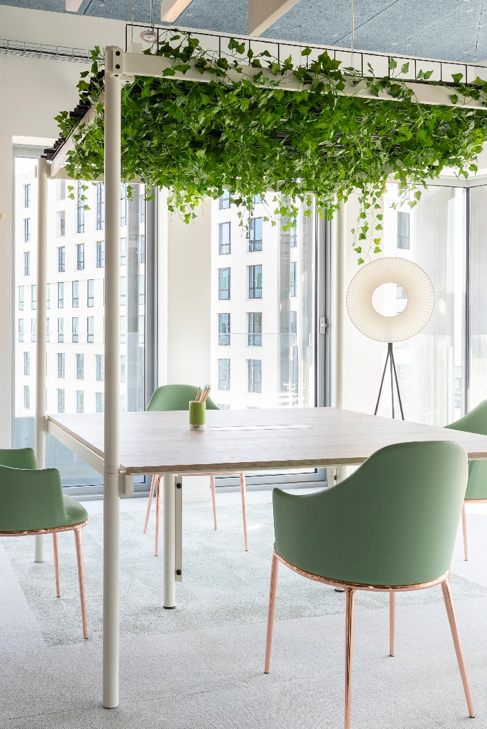 Meeting table with green chairs under a plant-covered pergola by large windows