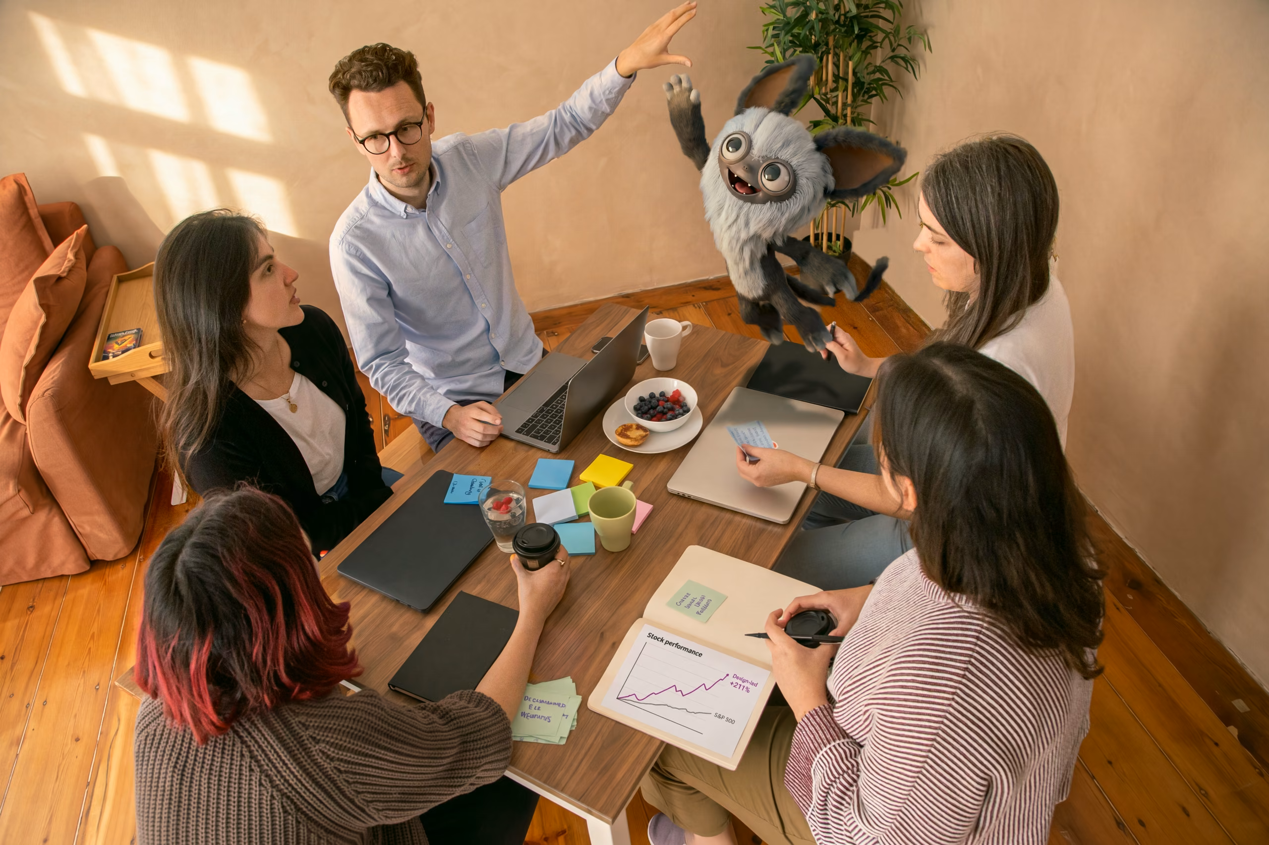Warm meeting room seen from above: a man raises his hand while presenting to four colleagues around a walnut table; Zap, the pale grey furry monster, has leapt off the table and flies mid-air toward the raised hand, sure it is a high five, while the printed Stock performance chart on the table shows design-led companies at +211% against the S&P 500.
