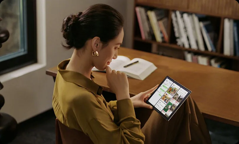 A woman in a mustard shirt sitting at a timber desk, reading a tablet in front of a bookshelf