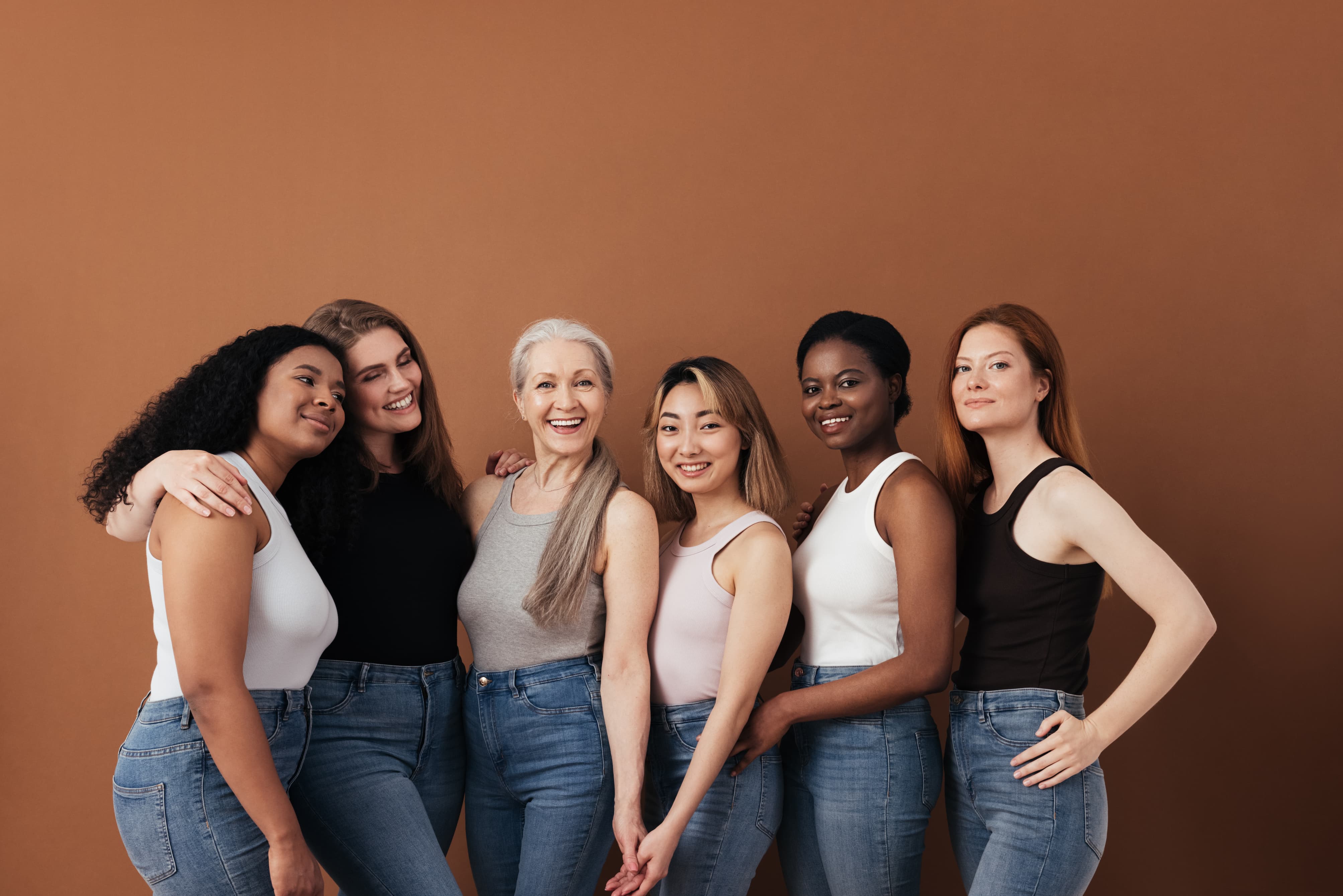 Portrait of six diverse women standing close together against a plain brown background, all wearing sleeveless tops and blue jeans, smiling and showing camaraderie.