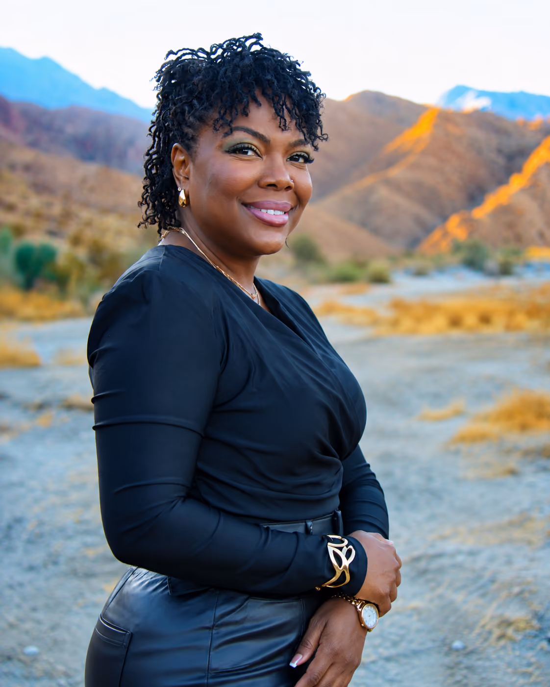 Smiling woman with curly hair wearing a black top and leather pants standing outdoors with mountains in the background at sunset.