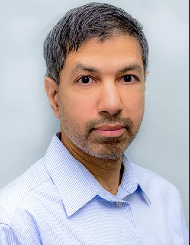 Portrait of a middle-aged man with short graying hair and a trimmed beard, wearing a light blue striped collared shirt, against a plain light gray background.