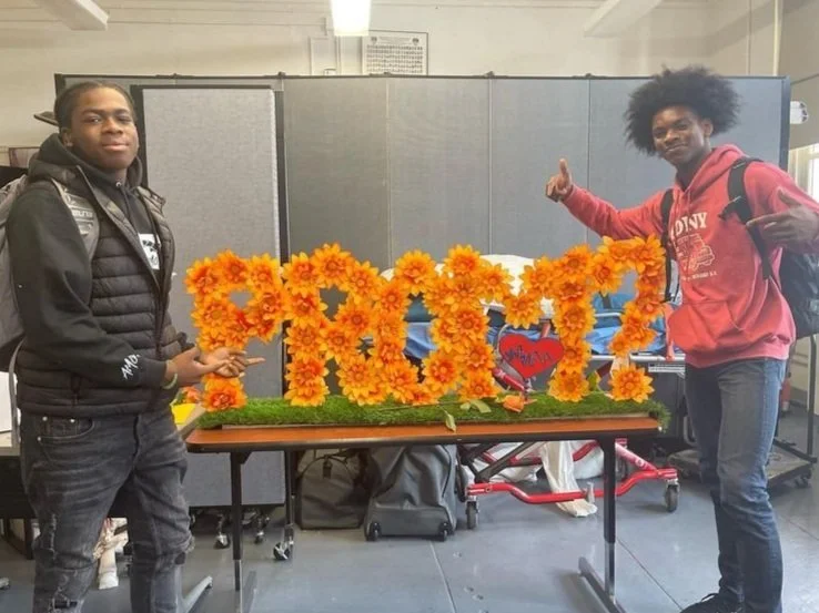 Two students posing with a floral prom proposal display on a table