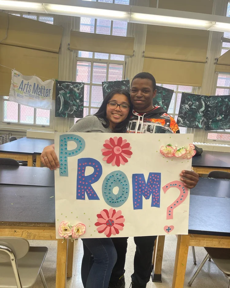 Two students holding a decorated prom proposal sign in classroom