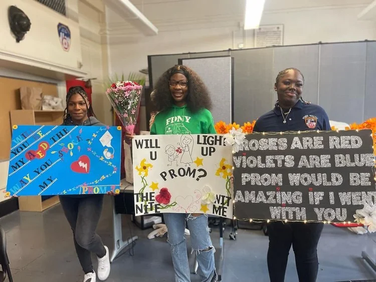 Three students holding handmade prom proposal signs at school