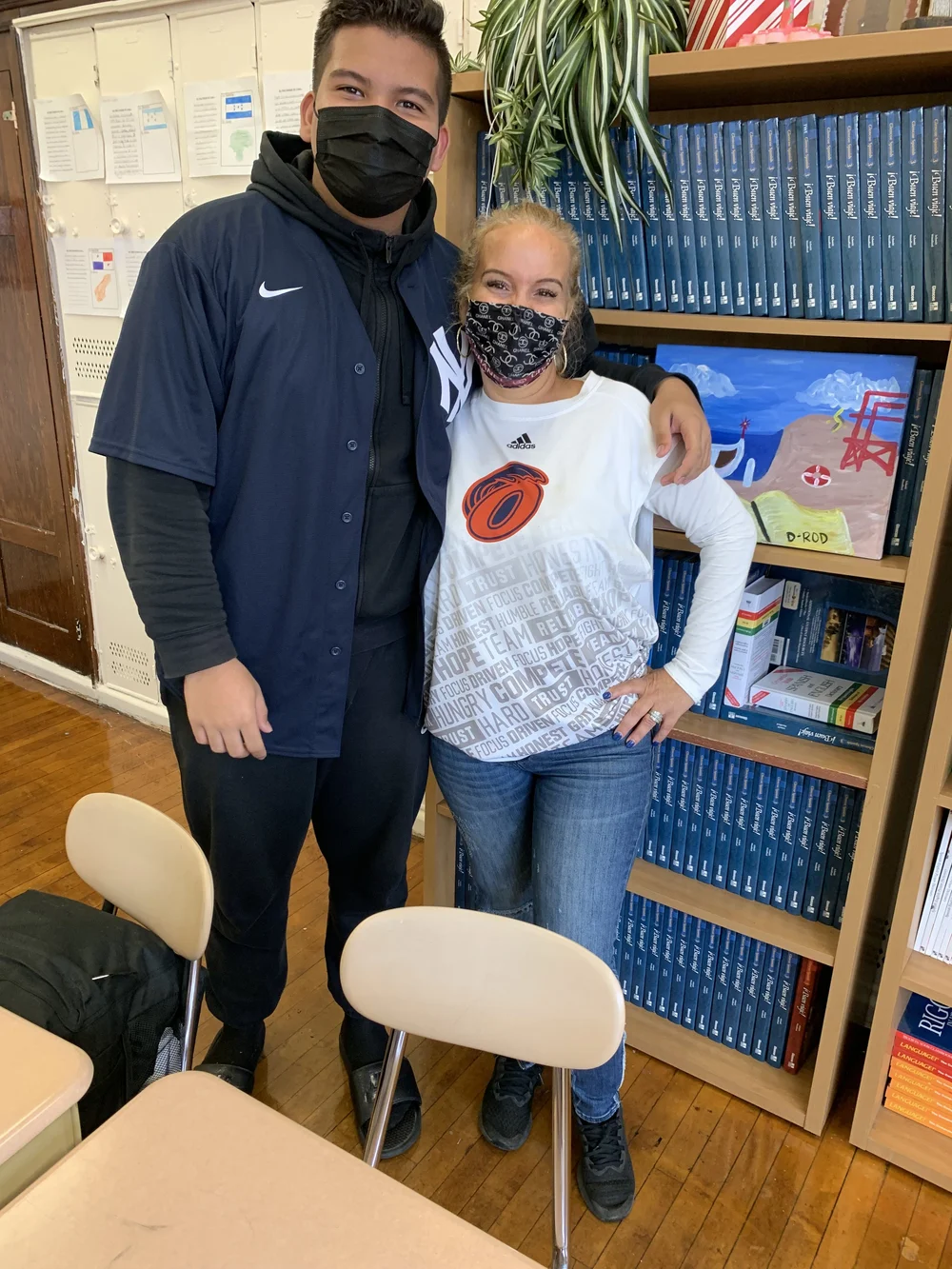 Two masked students posing together in a classroom near bookshelves