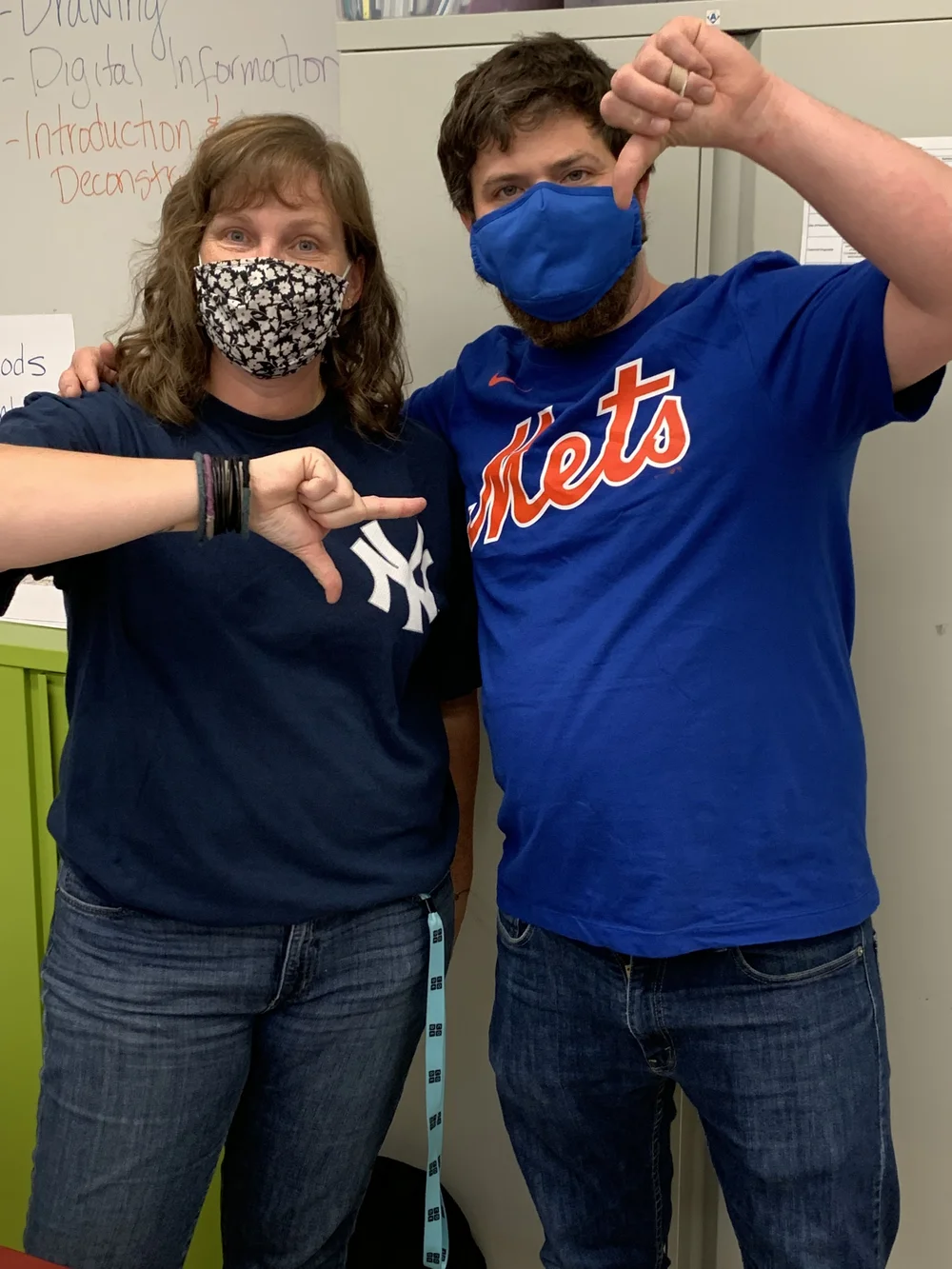 Two masked teachers giving thumbs down wearing rival Yankees and Mets shirts